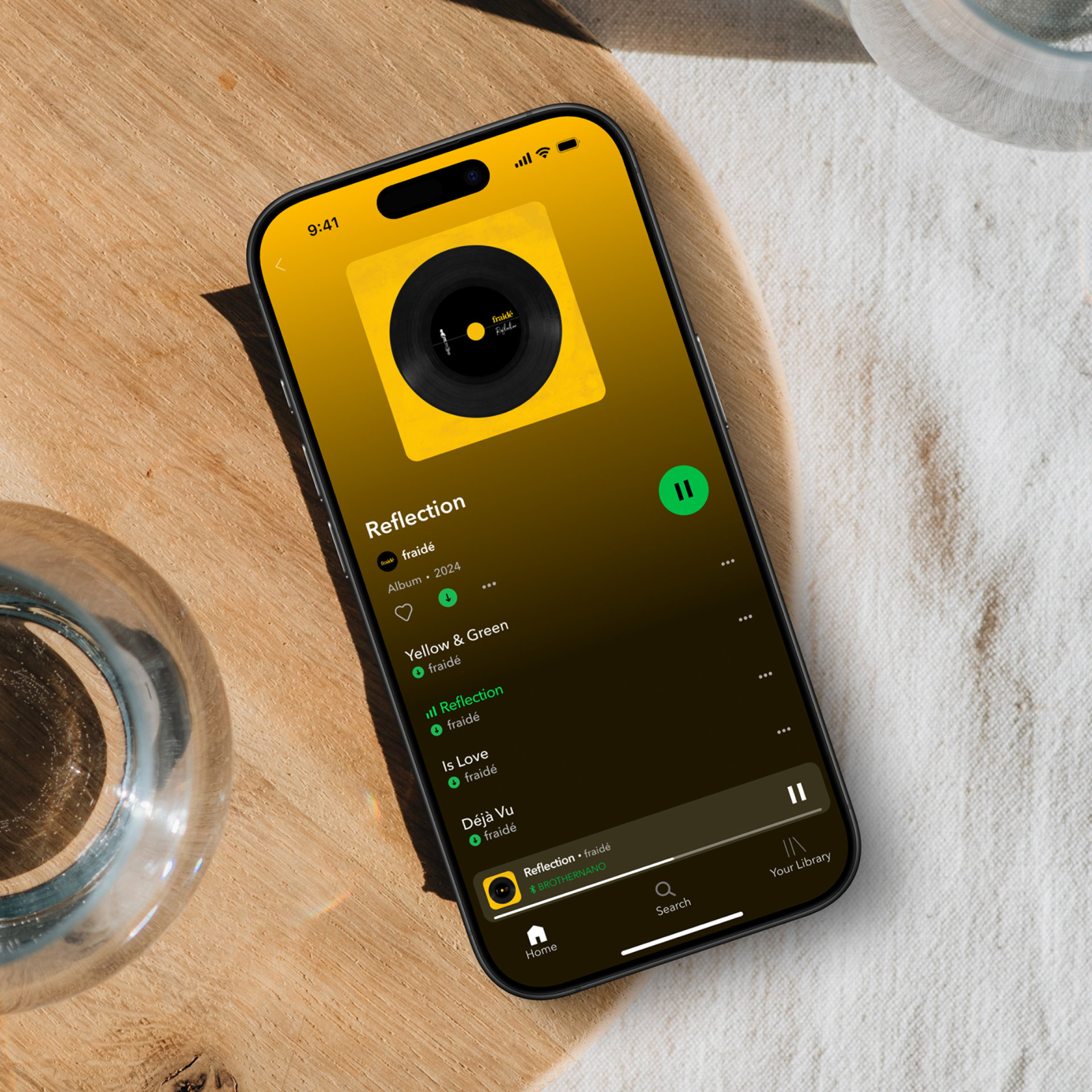 A smartphone on a wooden surface displays a music streaming app playing the song Reflection by Tokió, with a yellow album cover and playback controls visible. A glass of water is nearby.