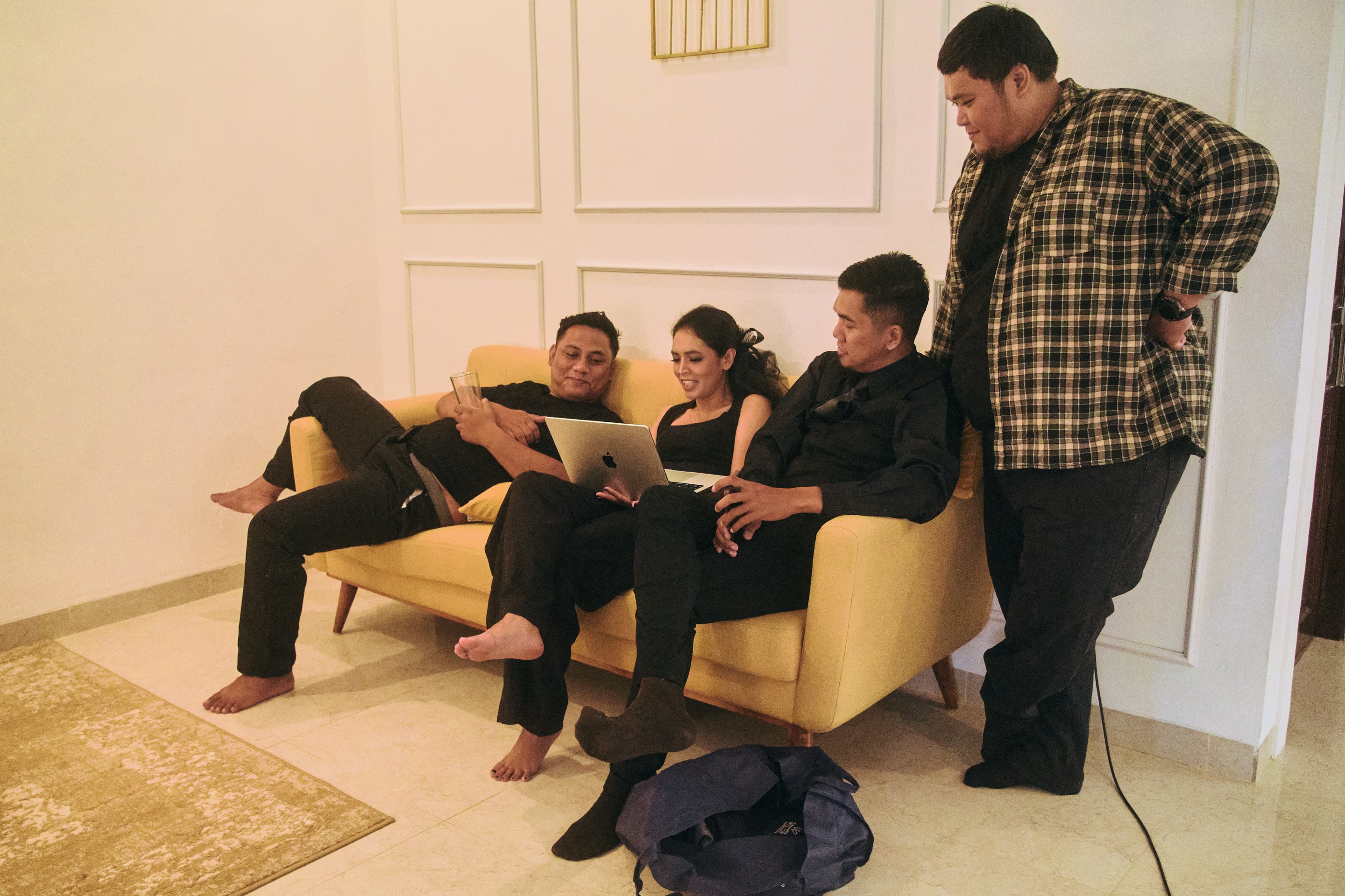 Four people sit and stand around a yellow couch, looking at a laptop together and smiling. The setting appears to be a cozy living room with light-colored walls and floor.