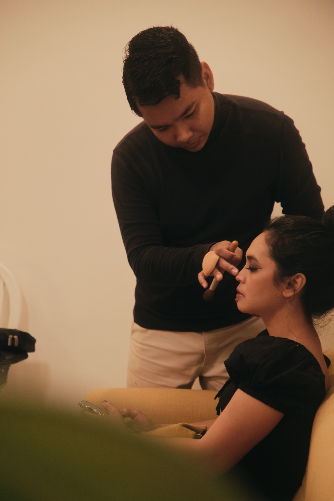 A person is sitting on a yellow chair while another person stands beside them, applying makeup to their face. The seated person holds a phone and looks relaxed, while the room has soft, warm lighting.