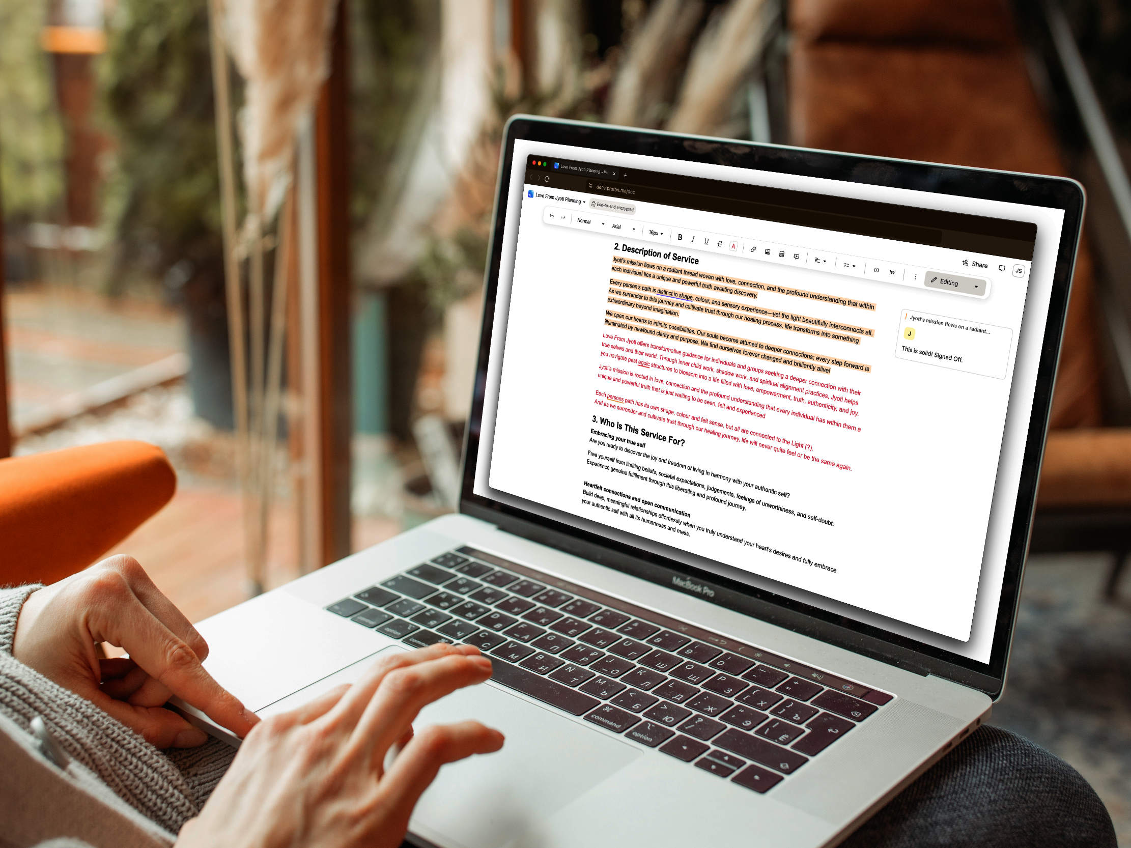 Person typing on a laptop, editing a document in Google Docs with highlighted text. The laptop rests on their lap in a cozy, indoor setting with plants and wooden decor visible in the background.